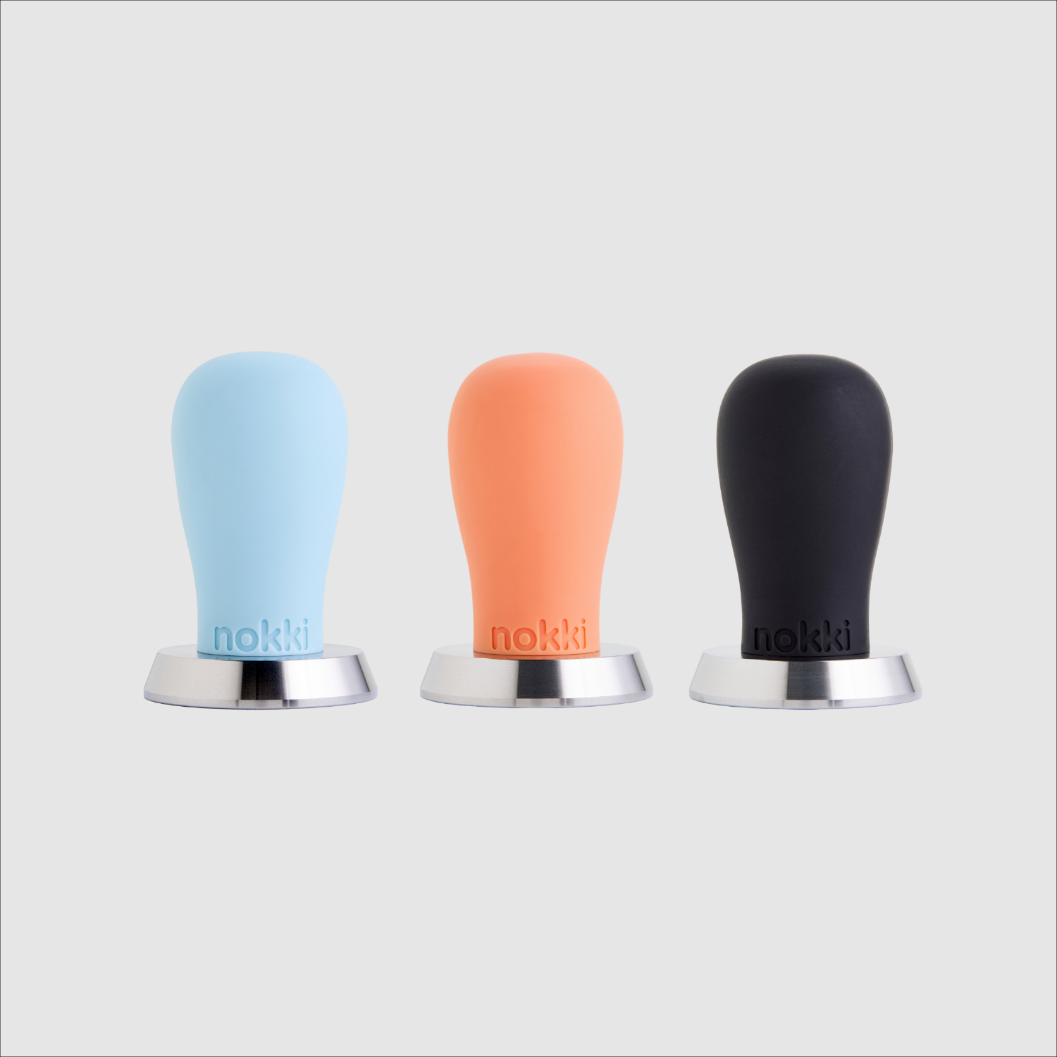 Three Nokki coffee tampers 51mm with matte silicone handles in light blue, orange, and black, each with a machined 316 stainless steel base, arranged side-by-side on a light gray background.